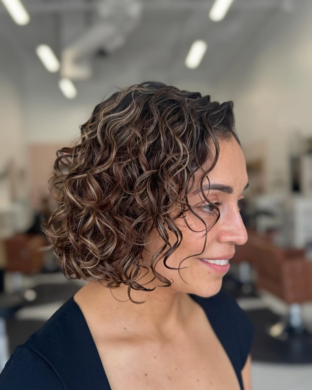Side profile of a smiling woman with a short, curly dark brown and blonde-highlighted bob with defined ringlets.