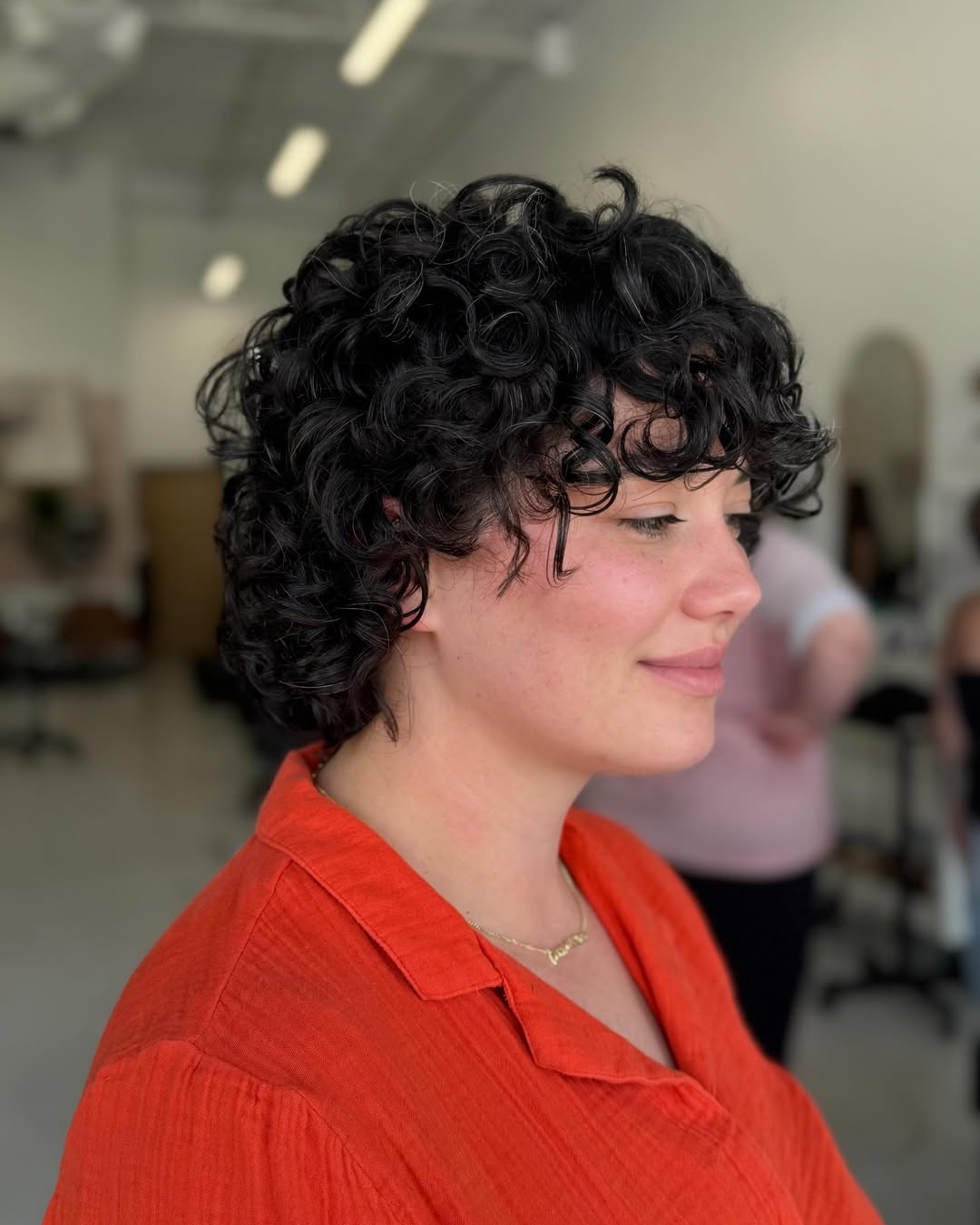 Side profile of a short, curly dark brown bob haircut with textured ringlets and bangs by Zoe.