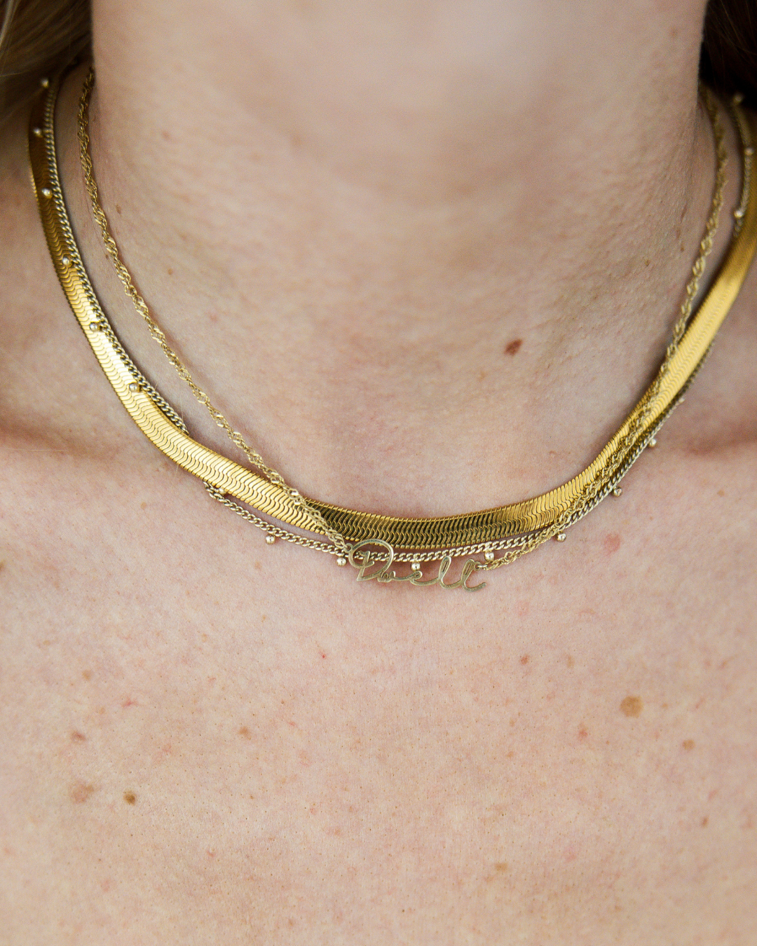 Close-up of layered gold necklaces, featuring a custom pendant with the Dwell Studio brand name.