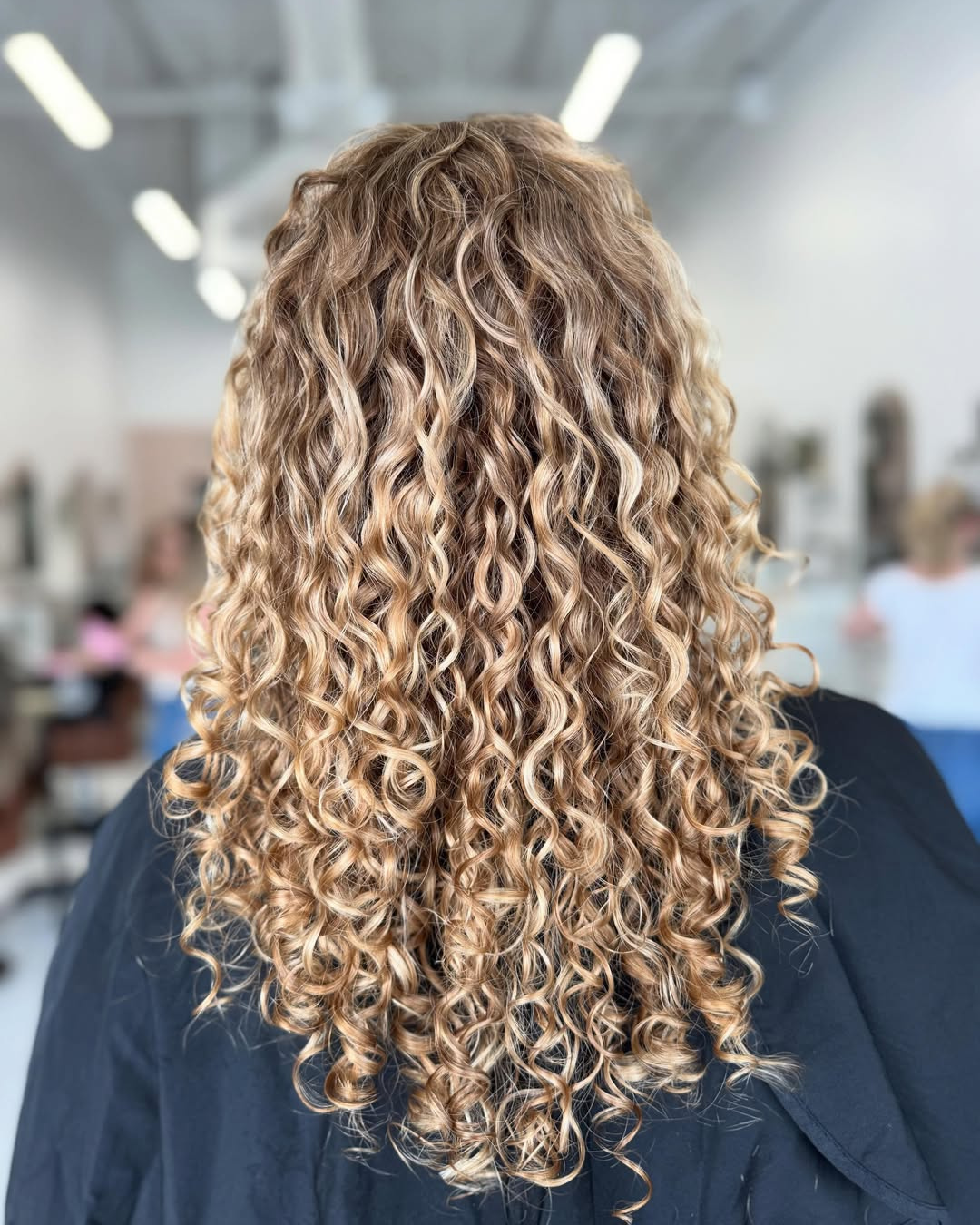 Back view of long, honey-blonde curly hair with tight, defined ringlets and dimensional highlights.