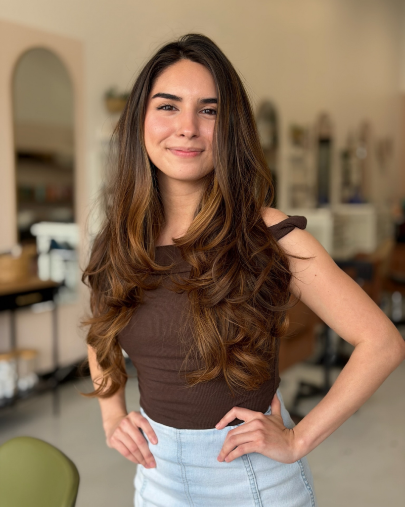 Stylist assistant Jackie Jasso with long wavy brown hair posing in Fort Worth salon.
