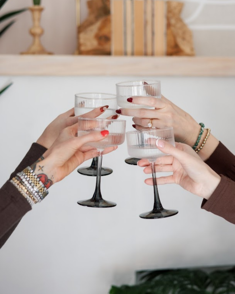 Four women toasting with ribbed glass coupe cocktails inside a bright hair salon