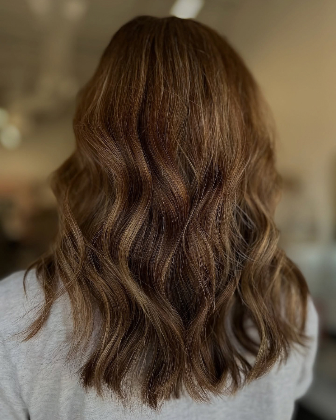 Back view of medium wavy brown hair with modern gray blending highlights by Ashley.