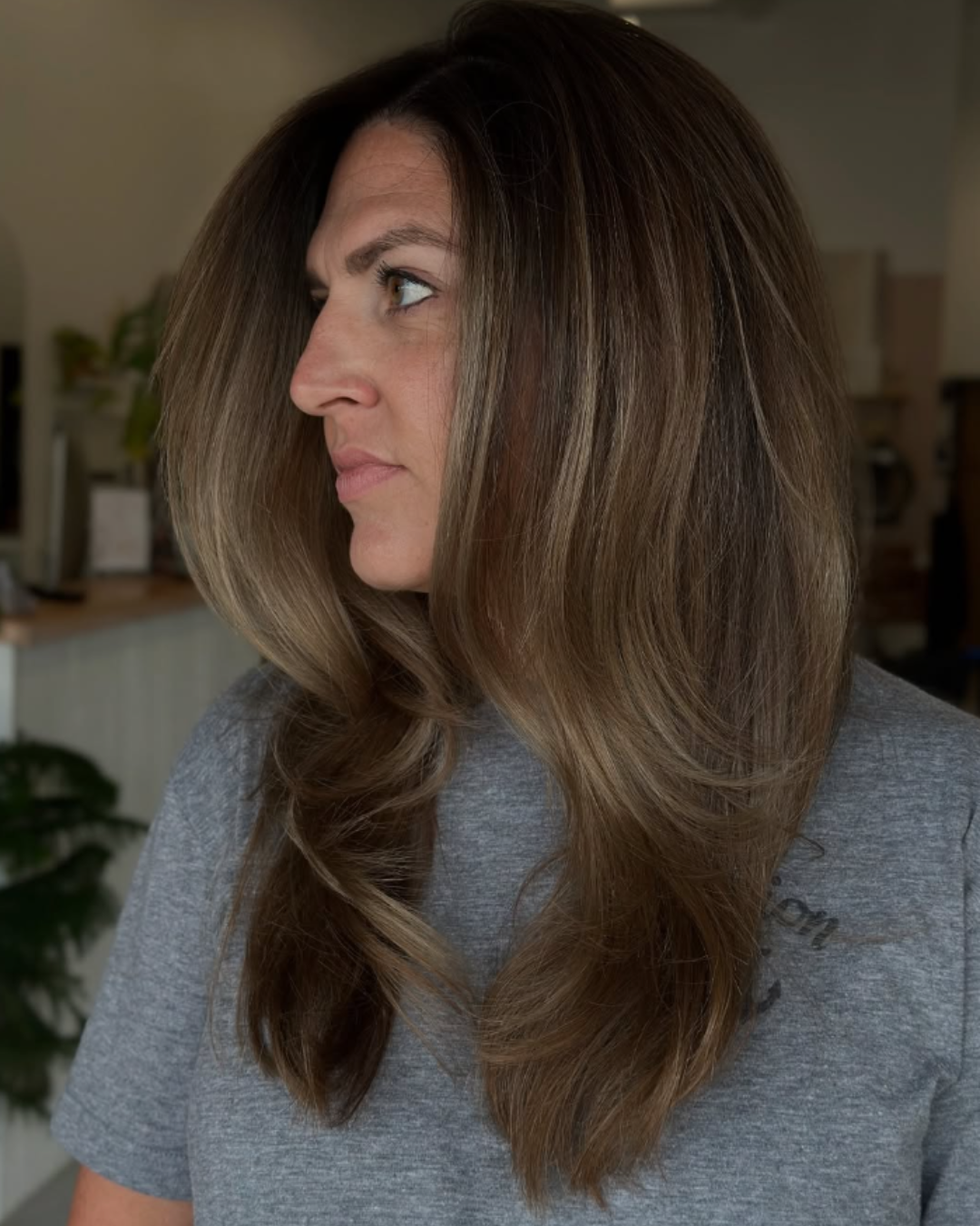 Woman with long, sleek ash brown layered hair styled with a fresh root touch-up and glossy finish by Angela.